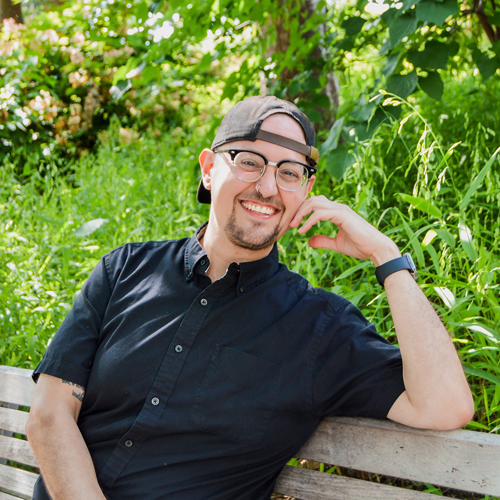 A headshot of Zach, in a black collared shirt and backwards black baseball cap, sitting on a bench in front of greenery. 